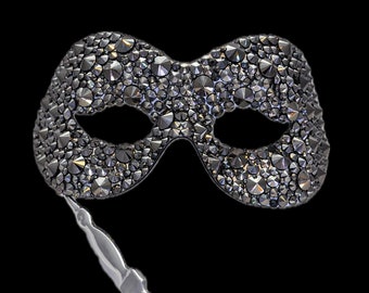Può includere: Una maschera da ballo nera ricoperta di strass. La maschera ha una forma tradizionale dell'occhio ed è attaccata a un lungo bastone sottile.