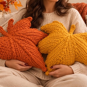 Può includere: Due cuscini a forma di foglia, uno di colore arancione vivace e l'altro giallo senape intenso. Entrambi i cuscini presentano un motivo a maglia uncinetto testurizzato con una vena centrale prominente e lobi appuntiti, che imitano il fogliame autunnale.