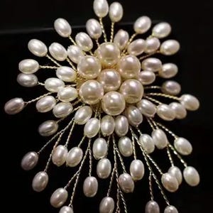 May include: A pearl brooch in a floral arrangement. The brooch has a central cluster of large, round pearls, encircled by smaller, oval pearls on fine gold-coloured wire stems. The background is black.