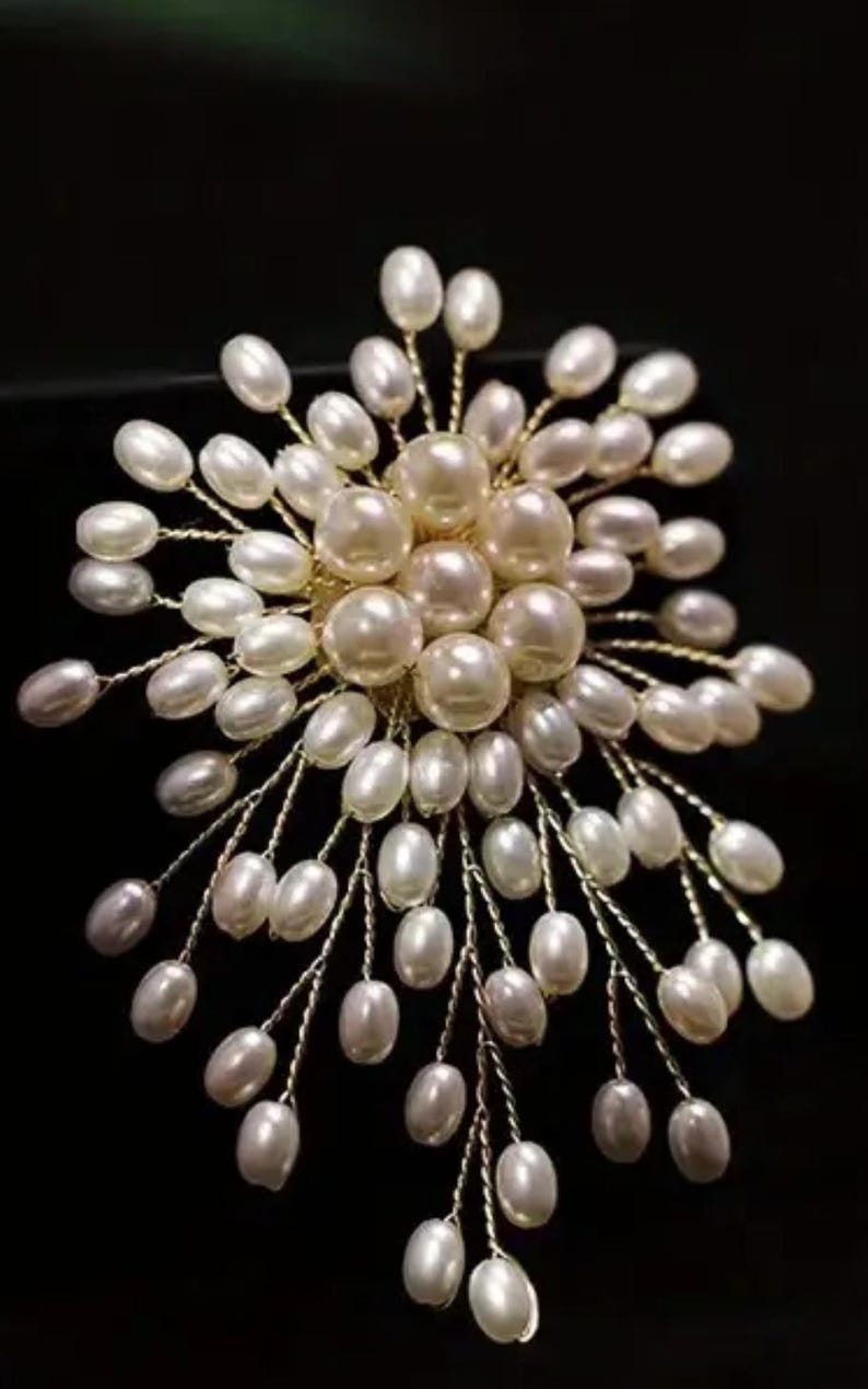 Puede incluir: Un broche de perlas con dise&ntilde;o floral. El broche presenta un grupo de perlas grandes y redondas en el centro, rodeadas de perlas ovaladas m&aacute;s peque&ntilde;as sobre delicados tallos de alambre dorado. El fondo es negro.