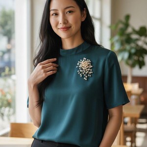 Puede incluir: Blusa color turquesa con cuello alto y mangas cortas. Un broche de perlas y dorado adorna el frente. La persona lleva pantalones negros. El fondo presenta una ventana y mesas de madera.