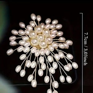 Puede incluir: Accesorio para el cabello con forma de flor de perlas blancas, de aproximadamente 7,7 cm de largo. El dise&ntilde;o presenta numerosas perlas blancas agrupadas en delicados tallos de alambre dorado, creando un arreglo floral. El fondo es negro.