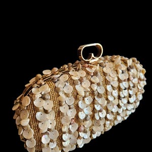 May include: A gold-toned clutch bag with a rounded rectangular form. The surface is adorned with small, round, off-white shell-like decorations and gold beads. A gold handle is fixed to the top.