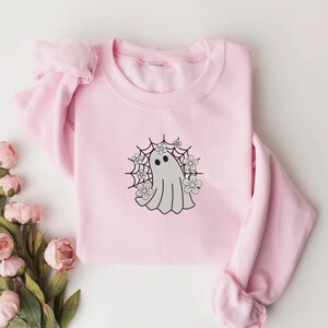 May include: A light pink sweatshirt featuring a gray ghost design surrounded by a spiderweb and flowers. The ghost has two black dot eyes. The sweatshirt is made of a soft material and is perfect for Halloween or everyday wear.