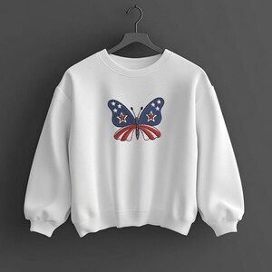 Puede incluir: Sudadera blanca con un dise&ntilde;o de mariposa bordada. La mariposa tiene alas azules con estrellas blancas y un cuerpo a rayas rojas y blancas. La sudadera est&aacute; colgada en una percha negra sobre un fondo oscuro.