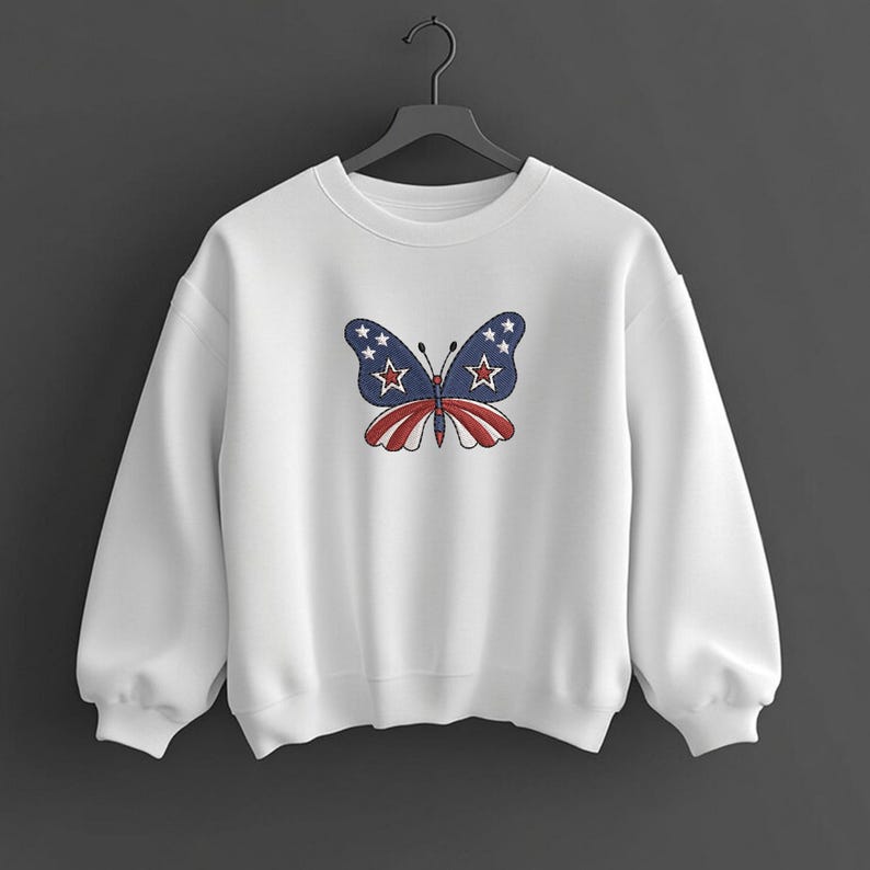 Puede incluir: Sudadera blanca con un dise&ntilde;o de mariposa bordada. La mariposa tiene alas azules con estrellas blancas y un cuerpo a rayas rojas y blancas. La sudadera est&aacute; colgada en una percha negra sobre un fondo oscuro.