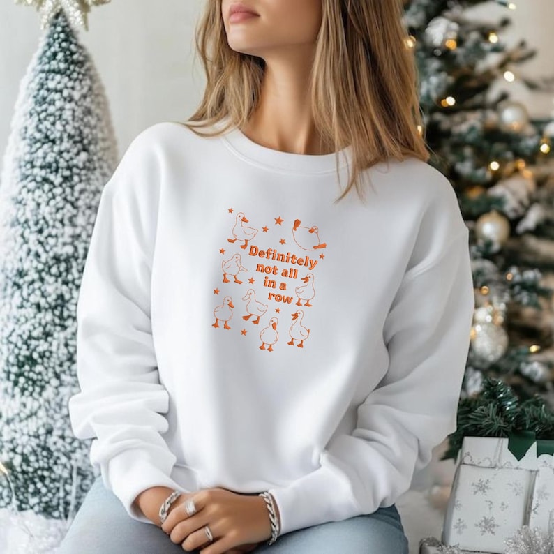 May include: White sweatshirt with an orange graphic of ducks and the text "Definitely not all in a row." The design includes stars and is centered on the front of the sweatshirt. The sweatshirt is a crew neck style.