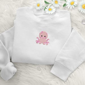 May include: White sweatshirt featuring a pink embroidered octopus design. The octopus has a smiling face and eight tentacles. The sweatshirt is made of a soft material and is suitable for casual wear.