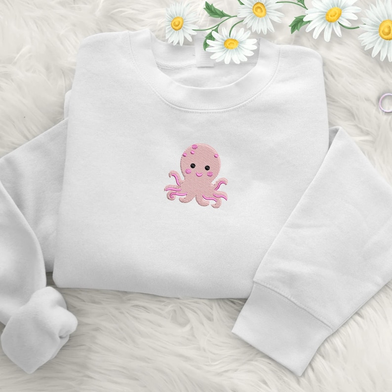 May include: White sweatshirt featuring a pink embroidered octopus design. The octopus has a smiling face and eight tentacles. The sweatshirt is made of a soft material and is suitable for casual wear.