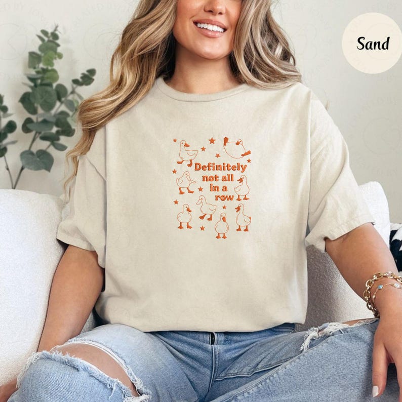 May include: Sand-colored t-shirt with a playful design featuring orange ducks and stars. The text "Definitely not all in a row" is printed in orange. The shirt is a relaxed fit.