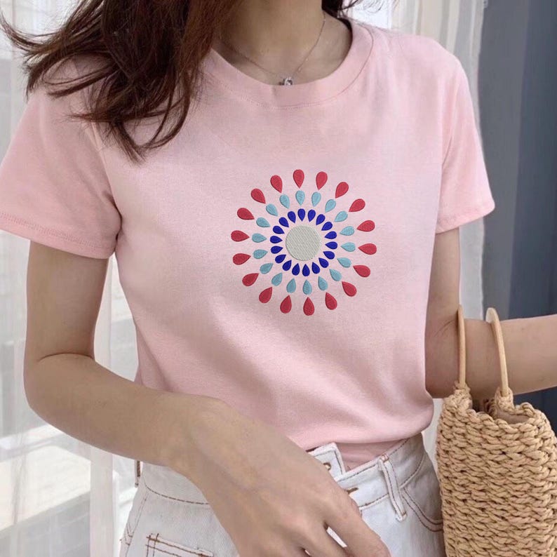 Puede incluir: Una camiseta rosa claro con un dise&ntilde;o circular de formas de l&aacute;grima rojas, azules y blancas. El dise&ntilde;o est&aacute; centrado en el pecho. La camiseta tiene mangas cortas y cuello redondo.