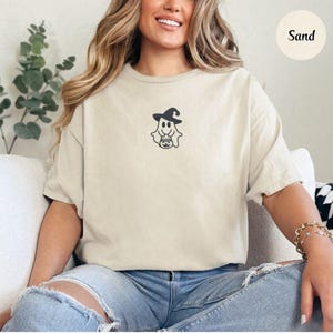 May include: Sand-colored t-shirt featuring a black and white cartoon ghost wearing a witch hat and holding a jack-o'-lantern. The shirt is a relaxed fit and has short sleeves. The word "Sand" is printed in a circle in the upper right corner.
