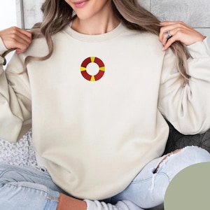 May include: Cream-colored sweatshirt featuring a red and yellow life preserver design on the chest. The sweatshirt has a crew neck and long sleeves. The life preserver is a classic nautical symbol.