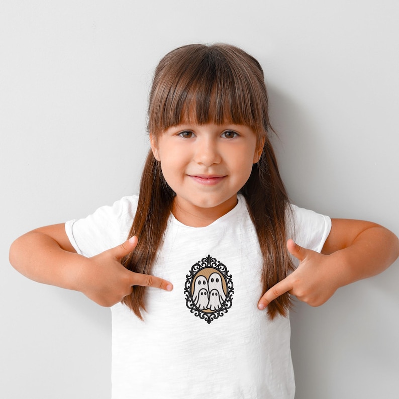 May include: White t-shirt featuring a decorative oval frame with two cartoon ghosts. The frame has a black and gold filigree design. The ghosts are white with black eyes and are set against a tan background.