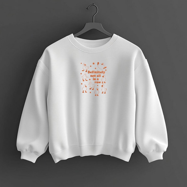 May include: White sweatshirt with the text "Definitely not all in a row" in orange, along with orange illustrations of geese and stars. The sweatshirt has a crew neck and long sleeves.