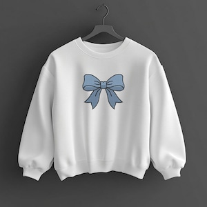 May include: A white long-sleeve crewneck sweatshirt featuring a centered graphic of a light blue bow with a dark blue outline. The sweatshirt has a ribbed collar, cuffs, and hem, and is displayed on a black plastic hanger against a dark background.