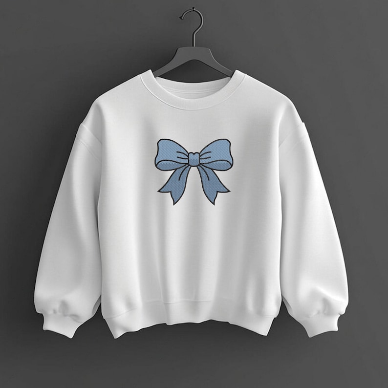 May include: A white long-sleeve crewneck sweatshirt featuring a centered graphic of a light blue bow with a dark blue outline. The sweatshirt has a ribbed collar, cuffs, and hem, and is displayed on a black plastic hanger against a dark background.