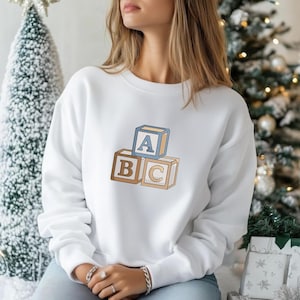 May include: A white long-sleeve sweatshirt featuring an embroidered graphic of three stacked wooden alphabet blocks. The top block is light blue with the letter A, while the two bottom blocks are tan with the letters B and C respectively.