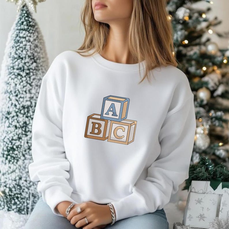 May include: A white long-sleeve sweatshirt featuring an embroidered graphic of three stacked wooden alphabet blocks. The top block is light blue with the letter A, while the two bottom blocks are tan with the letters B and C respectively.