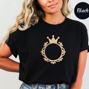May include: A black short-sleeved t-shirt featuring a centered gold-colored embroidery design. The graphic consists of a circular ornate frame with decorative scrollwork details, topped with a small crown icon. The shirt has a crew neckline.