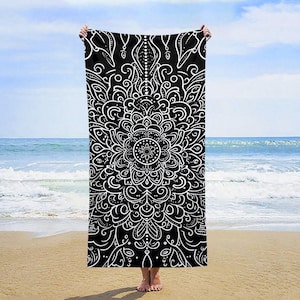 May include: Black and white beach towel featuring a mandala design. The rectangular towel is held up against a beach and ocean backdrop. The intricate white pattern contrasts against the black background, creating a striking visual.