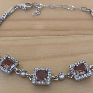 May include: A silver bracelet with three square, peach-coloured gemstones framed by small, clear stones. The bracelet has a delicate chain design and is shown on a light brown wooden surface.