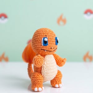 May include: A handmade crochet plush toy of the character Charmander, featuring a bright orange body, a cream-coloured belly, blue eyes, and a small flame at the tip of its tail. The figure has white claws on its feet and a stitched mouth.