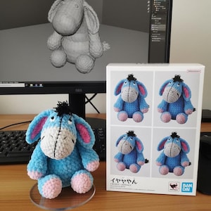 May include: A crocheted donkey plush toy in shades of blue, gray, pink, and white, with a black tuft of hair. The toy is displayed on a clear stand, next to a box featuring the same design. The box has Japanese text.