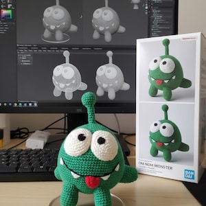 May include: A green crocheted Om Nom Monster toy with white eyes, a red tongue, and a long antenna. The toy is displayed on a clear stand. A box with the same toy is visible, along with a computer screen showing 3D model designs.