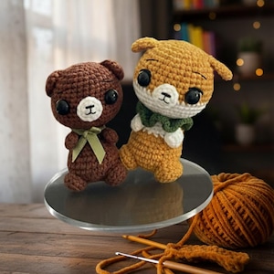 May include: Two handmade crochet stuffed animals: a brown bear with a green ribbon and a tan fox with a green scarf. They sit on a clear, round surface. A ball of orange yarn, crochet hooks, and a wooden table complete the scene.