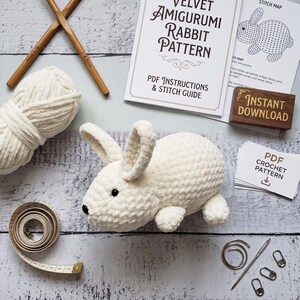May include: A white velvet amigurumi rabbit, a ball of white yarn, two wooden crochet hooks, a measuring tape, a metal needle, stitch markers, and printed pattern instructions titled Velvet Amigurumi Rabbit Pattern with an Instant Download label.
