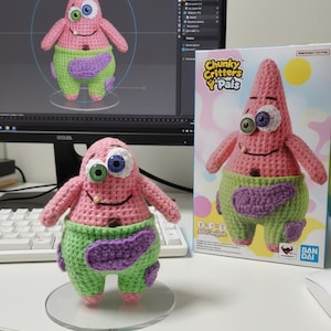 Może przedstawiać: Ręcznie robiona szydełkowa pluszowa zabawka Patrick Star, z różowym korpusem, zielonymi szortami i fioletowymi akcentami. Zabawka ma jedno niebieskie i jedno zielone oko. W tle pudełko z napisem "Chunky Critters Y' Pals".