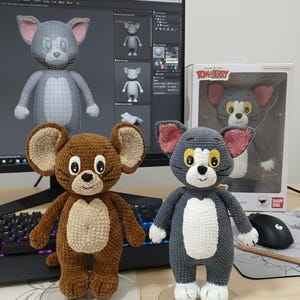 May include: Two crocheted Tom and Jerry figures. One is brown, resembling Jerry, and the other is gray, resembling Tom. Both have large ears with pink inner ears, white bellies, and are standing on clear stands. A computer screen displays a 3D model of Tom.