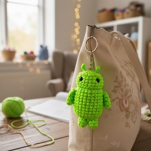 Op de afbeelding: Een felgroene, gehaakte alien-sleutelhanger hangt aan een beige draagtas met bloemenborduurwerk. De alien heeft grote zwarte ogen en kleine ledematen. Een bol groen garen ligt op een houten tafel, wat op handwerk duidt.
