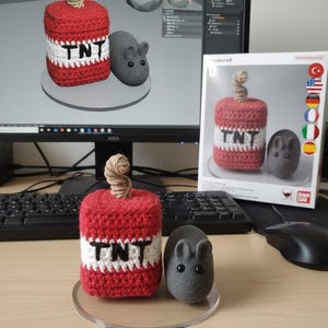 May include: A red and white crocheted TNT dynamite with the word "TNT" and a gray mouse figurine. The dynamite has a twisted brown fuse. The mouse has small ears and black eyes. The items are on a clear circular stand.