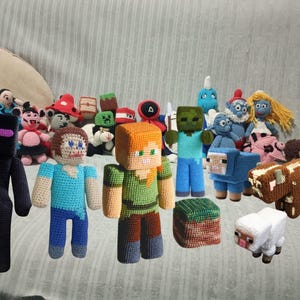 Könnte beinhalten: Eine Sammlung handgefertigter Häkelpuppen und -figuren, darunter Charaktere aus Minecraft und anderen Fantasy-Themen. Die Puppen weisen leuchtende Farben und detaillierte Nähte auf, mit verschiedenen Größen und Designs. Die Sammlung umfasst einen schwarzen Enderman, Steve, Alex, einen Zombie und ein Schaf.
