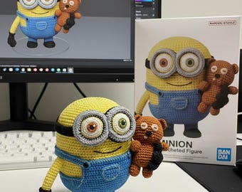 Peut inclure: Une figurine Minion au crochet, jaune et bleu, tenant un petit ours en peluche marron. Le Minion a de grands yeux ronds et des gants noirs. Une boîte avec une image similaire est en arrière-plan.