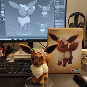 May include: A crocheted Eevee figure with brown, white, and pink accents, wearing a floral crown. The figure is displayed on a clear stand, next to a box with a similar image. A computer screen shows a 3D model of the figure.