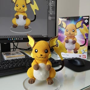 Könnte beinhalten: Gehäkelte Raichu-Figur in Gelb, Braun und Weiß. Das Plüschtier hat einen runden Körper, spitze Ohren und einen Blitzschwanz. Es wird auf einem transparenten Ständer ausgestellt, mit einem Computermonitor und einer Box mit der Raichu-Figur im Hintergrund.
