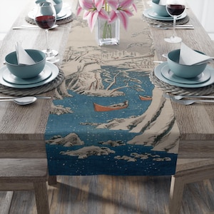 May include: A table runner with a Japanese-inspired landscape design. The runner features a blue river scene with orange boats, snow-capped mountains, and trees. The runner is set on a wooden table with place settings, wine glasses, and a vase of pink flowers.