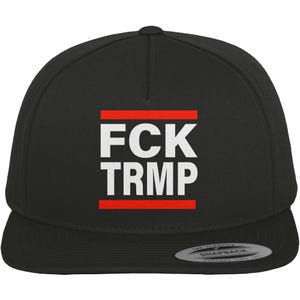 Könnte beinhalten: Schwarze Snapback-Kappe mit dem weißen Text "FCK TRMP", der von zwei roten Linien eingerahmt wird. Die Kappe hat einen flachen Schirm und einen Schnappverschluss auf der Rückseite. Der Markenname "Snapback" ist sichtbar.