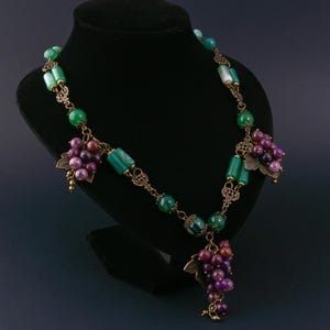 Peut inclure: Un collier avec des perles vertes et violettes, et des pendentifs en forme de grappes de raisin de couleur bronze. Le collier est exposé sur un mannequin noir. Les perles sont de formes variées, rondes et rectangulaires. Les grappes ont des feuilles en bronze.