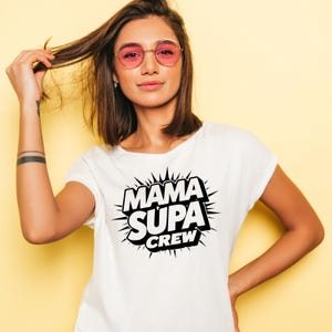 May include: White t-shirt with the text "MAMA SUPA CREW" in a black, comic-style design. The shirt is worn by a person with pink-tinted glasses and dark hair. The background is a solid yellow.