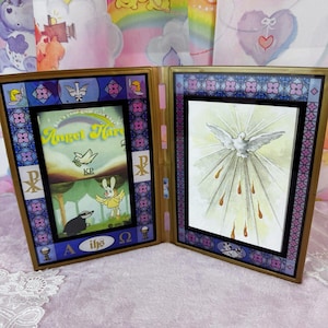 May include: A two-panel hinged picture frame with gold trim. The left panel features a cartoon scene with a rabbit and badger, and the text "Angel Hare." The right panel displays a dove with rays of light.