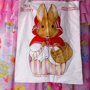 May include: A white fabric panel featuring Mrs. Bunny from Beatrix Potter's Nursery Classics. The illustration shows a brown rabbit in a red hooded cloak, holding a basket and a handkerchief. The panel has dotted edges and text that reads "LEAVE OPEN FOR STUFFING."