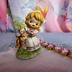 May include: A ceramic figurine of a blonde child wearing a white bonnet and dress with pink accents, standing next to a small brown fox on a green base. Beside the figurine lies a gold-toned bracelet featuring rectangular pink faceted crystals.