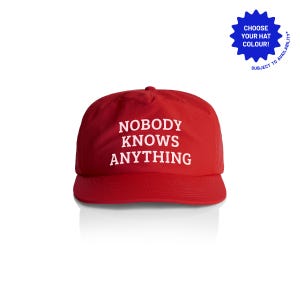 May include: A red baseball cap with the phrase "NOBODY KNOWS ANYTHING" in white block letters. The hat has a curved brim and a classic six-panel design. A blue starburst graphic is in the upper right corner.