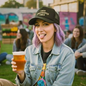 May include: A black trucker hat with the text "I MISS PRECEDENTED TIMES" in white. The hat is worn by a person with purple hair, a denim jacket, and a beer in hand. The background shows a park setting with other people.