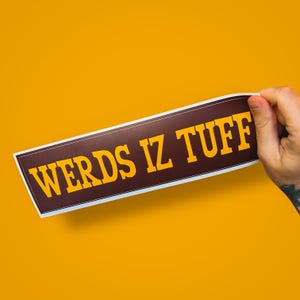 May include: A rectangular sticker with the text "WERDS IZ TUFF" in yellow letters on a brown background. The sticker is held by a hand against a bright orange background.