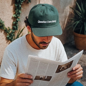 Puede incluir: Gorra de béisbol verde oscuro con el texto blanco "Question Everything". Una persona lee un periódico, usando la gorra, una camiseta blanca y vaqueros azules.